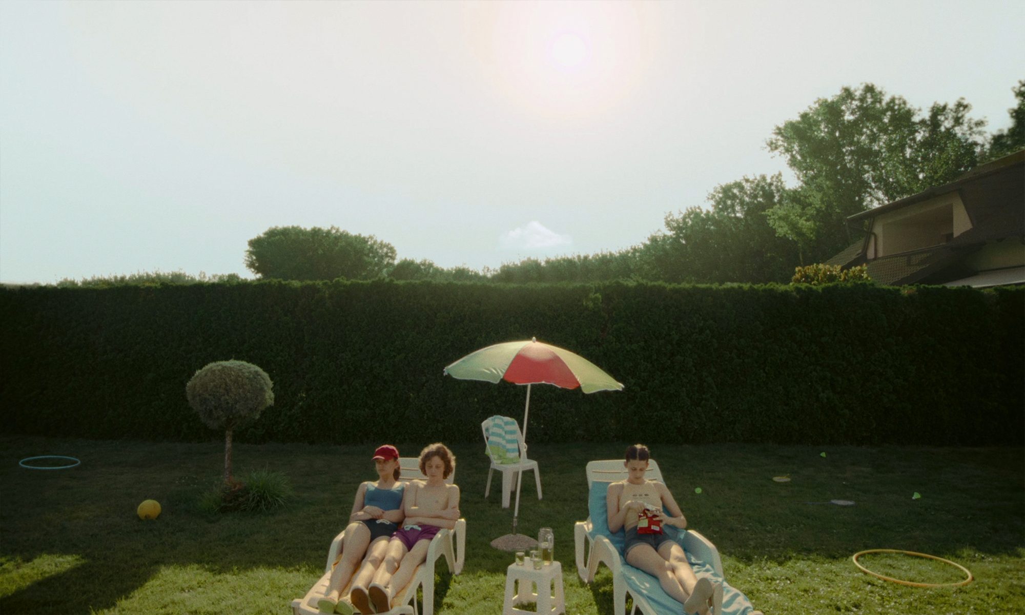 Three young people on sun loungers in a garden under a parasol